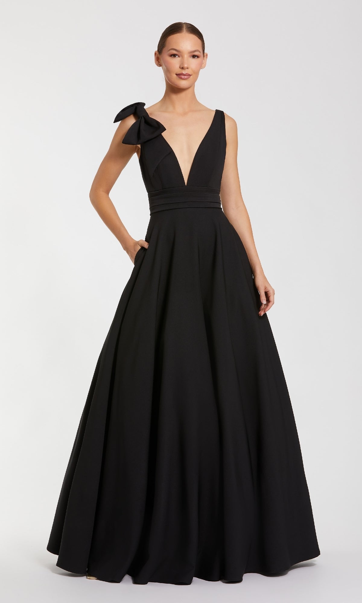A woman in the Mac Duggal 49972 Shoulder-Bow Long Formal Dress, featuring a sleeveless floor-length design with a deep V-neckline and a statement bow on one shoulder, stands against a plain background.